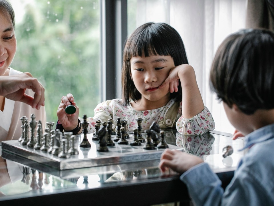 Two children playing chess while a grandparent watches